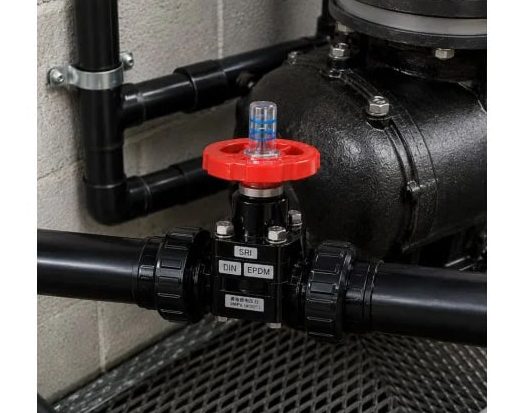 Industrial shut-off valve with a red handwheel on a black pipe network beside a large pump and concrete wall.