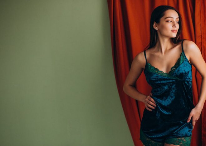 Woman in teal velvet lingerie posing against a green wall with red curtains, hands on hips and looking off to the side.