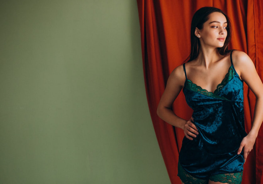 Woman in teal velvet lingerie posing against a green wall with red curtains, hands on hips and looking off to the side.